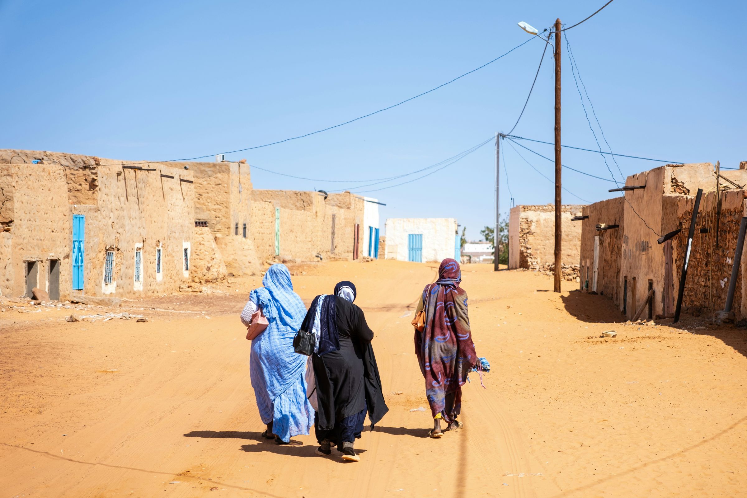 Three women walking together down a sandy street lined with adobe buildings.