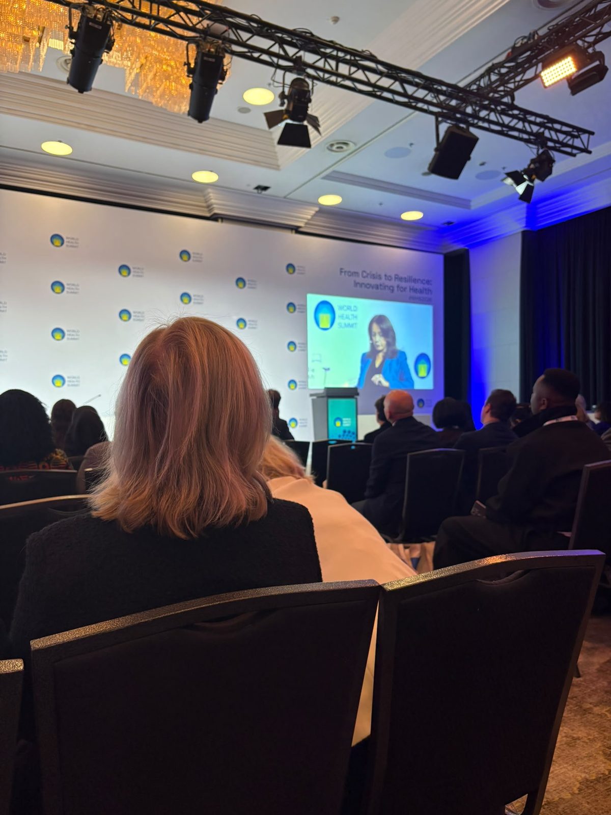 A speaker on screen in a plenary hall under the summit theme From Crisis to Resilience: Innovating for Health.